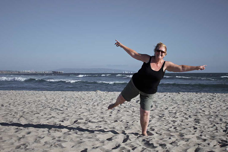 Félix Ossa - Dónde está Colombia? - Imagen de una mujer en la playa. Su cuerpo está balanceado en una pierna y sus manos y otra pierna están apu